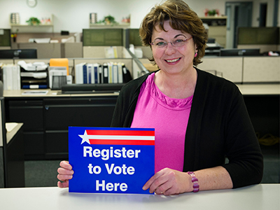 Elections staff member at counter holding "register to vote here" sign