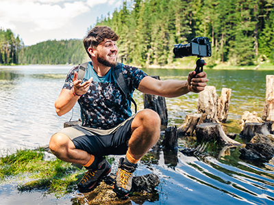Male students poses with video camera while squating on a log at the shore of a lake