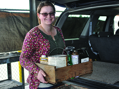 white woman standing next to open hatchback of a vehicle holding a wooden box of household cleaners 