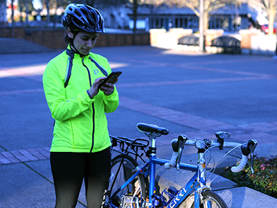 Woman in a bike helmet wearing a neon yellow windbreaker, stands next to her bike while looking at a map on her smartphone.