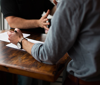 Close-up image of two men seated at a table. One man is gesturing with this hands.