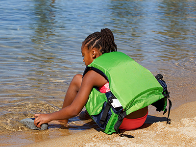Black female youth wearing bathing suite and lime green life vest, sits at the water's edge of a lake.