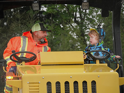 White male Public Works employee looks at white child while sitting in the cab of a tractor.