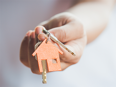 A person's hand is extended toward the camera. They are holding a key right containing a key and small wooden, orange house emblem.