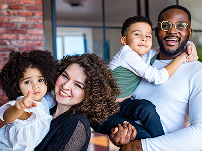 Mixed race family of two adults and 2 children smiling at the camera. Parents are each holding a child in their arms.