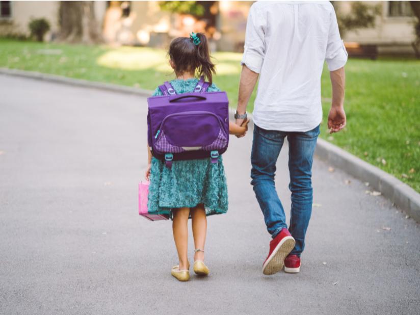 A young girl with a backpack being walked to school by her parent. 