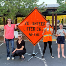 Two adults and two young people stand next to an orange sign that reads "Volunteer Litter Crew Ahead." 