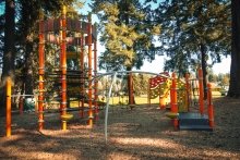 An orange playground with bark chip surface underneath sits under tall evergreen trees as sunlight streams through the tree canopy on a clear day.