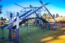 A purple and gray playground with rope ladders sits in the shade on a sunny, clear day.