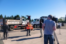 A worker wearing a reflective, florescent colored vest stands in front of road construction vehicles speaking with a reporter, with a videographer filming, on a cloudless day.