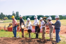Seven people wearing hardhats stand behind a dirt mound in an open field, using gold shovels to toss dirt. An orange construction barrier and forested hills are seen in the background. 