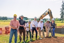 Seven people wearing hardhats stand behind a dirt mound in an open field, holding gold shovels. An orange construction barrier, excavator and forested hills are seen in the background. 