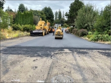 Two yellow construction vehicles are on fresh, smooth, dark black pavement. The pavement in the foreground is old, brown, worn and rough. A tree-lined residential neighborhood is in the background.