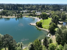 Aerial view of a blue-green pond with a sandy shore. An area of green grass and deciduous trees borders the pond.