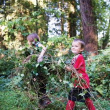 Children carry piles of weeds after removing them from a forested area.