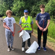 Two teens and one adult stand on a paved trail in a natural area, holding litter grabbing sticks and partially filled litter collection bags. 