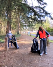 Two volunteers wearing masks pick up litter using litter grabbing sticks and trash bags, in a forested park.