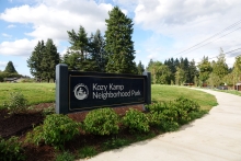A paved path curves to the right around a navy blue sign that says "Kozy Kamp Neighborhood Park". Low green plants grow in front of the sign and a green field, evergreen and deciduous trees are behind with a blue sky and clouds above.