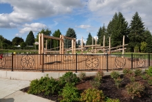 A playground with large wooden timbers is behind a metal fence with decorative designs. A paved path and landscaping are in front of the playground and trees, a blue sky and clouds are seen behind the playground.