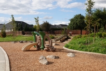 A playground sized for small children, with climbing rocks, a play structure and a slide built into a small hillside, is on a bark chip play surface. Landscaped beds are on either side of the playground. Homes and trees are visible in the background. A blue sky is dotted with clouds. 