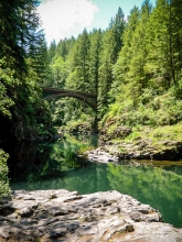 Beyond a grayish/brown rocky outcropping is an emerald green, smooth and still river. In the background, a large timber bridge high above the water connects two sides of a valley covered with evergreen trees. 