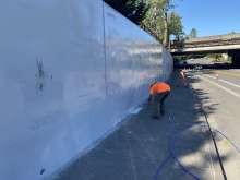 A wall approximately 10 feet tall curves from the left towards the center of the photo. The wall is shiny and a neutral color, showing the wetness of recently applied primer. A worker in a florescent orange shirt is standing on the sidewalk in front of the wall, bending over to paint a low area on the wall. A road is adjacent to the wall and sidewalk, on the right. A freeway overpass and trees are in the background. The sky is blue and cloudless.