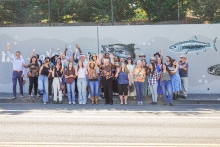 A group of several dozen people smile, wave and pose while standing on a sidewalk in front of a large retaining wall. The wall is gray and painted with gray, black and white fish. There is a chain link fence at the top of the wall and green vegetation behind the fence. It is a sunny day. 