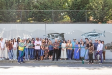A large group of dozens of people stand on a sidewalk between a road and retaining wall. The wall behind them is gray and is painted with gray, black and white fish. Above the wall is a chain-link fence, behind which is green vegetation. It is a sunny day. A woman in the middle smiles at the camera while she holds giant scissors, cutting a green ribbon. 