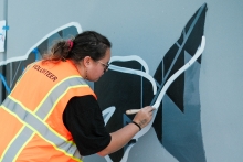 A person with long hair, wearing a florescent orange vest that says, "volunteer", uses a small brush to add white paint to a white, black, and gray fish on a gray retaining wall. 