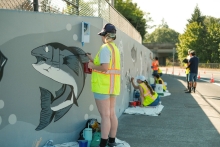 Volunteers wearing florescent yellow and orange vests paint gray, black and white fish on a gray retaining wall. One volunteer is standing in the foreground painting a fish, two volunteers in the background are sitting on the ground painting, and a volunteer seen in the far background is standing to paint. They are on a sidewalk in the shade on a sunny day. The retaining wall is topped with a chain link fence. There are green leafy trees in the background.