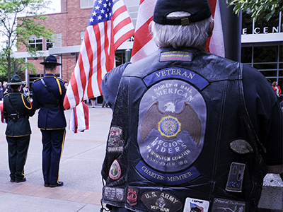White male veteran wearing a vest with patches, photographed from the back and standing at attention.
